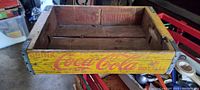Front side of wooden Coca Cola crate showing yellow paint with red Coca Cola logo and 'Drink Coca Cola in Bottles' text.