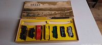 Open box showing vintage Champ train set in yellow tray with six train cars arranged neatly. Box top visible with scenic black-and-white illustration and 'CHAMP' branding.