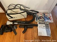 Full view showing Oreck XL compact canister vacuum, black hose, metal extension, and multiple attachment heads. Also shows unopened packs of vacuum bags and user manual.