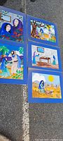 Five hand painted religious art pictures depicting Bible story scenes, laid out outdoors on pavement. Mounted on blue backing, bright bold watercolor paintings, approx 26" x 20" each.