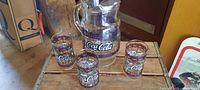 Glass pitcher and four glasses arranged on wood surface clearly showing Coca-Cola logos and Cactus Willys restaurant promotional branding from mid 1980s.
