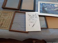 Photo of stitchery sampler dated 1834 alongside various framed prints on a grey quilted surface.