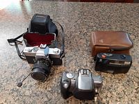 Minolta SR-T 303b camera with lens and strap alongside Kodak camera case and compact film camera in black with case on countertop.
