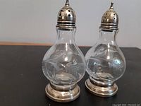 Pair of clear glass salt and pepper shakers with intricate etched designs, sterling silver bases and pierced caps, shown on black surface.