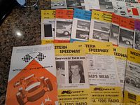 Assorted speedway programs laid out on a dark table, showing colorful covers with car racing images and text including 'Western Speedway Official Programme' and several dated programs from the 1970s.