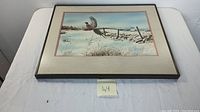 Photo showing framed watercolor painting with a bird (pheasant) in flight above snowy landscape with fence and trees in distance