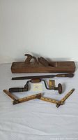 All tools displayed: wooden hand plane, hand drill, sharpening tool, folding ruler