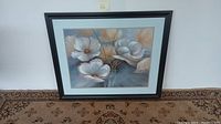 Framed floral oil painting showing large white and beige flowers with subtle shading, signed by the artist, placed on carpeted floor.
