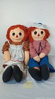 Front view of two large handmade plush Raggedy Ann style dolls seated side by side on a blue patterned cloth.