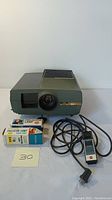 Front view of green Sawyer's Grand Prix 570 AF projector with lens, adjacent two boxed replacement lamps and wired remote control with power cord.