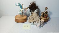 Four vintage collectible items including a porcelain doll, bisque baby figurine, white carousel horse music box, and an animal scene music box with rabbit and bear around lamppost.