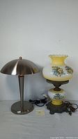 Pair of table lamps shown side by side on a white surface, including one modern metal lamp and one yellow floral hurricane lamp with metal base.