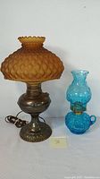 Both lamps shown side by side with a white background, clearly showing the amber frosted shade on electric metal lamp and blue glass oil lamp.