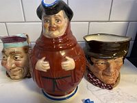 Three Toby Jugs placed side-by-side on a white tile background showing different facial expressions and clothing styles.
