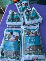 Five 8 lb bags of Kaytee Basic Nyjer Seed arranged on a purple surface, showing front of packaging with bird illustrations