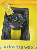 Close-up photo of Invicta men's wristwatch showing black dial with chronograph and yellow silicone strap inside the original watch compartment.
