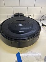 Front-facing view of black iRobot Roomba e5 on countertop with white subway tile background, showing domed top and CLEAN button.