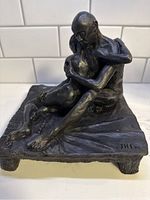 View of metal sculpture showing two seated figures embracing, front angle, signed JHT 98 on base