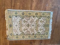 Full view of small hand knotted wool carpet showing central diamond and floral pattern with beige and green colors, fringed edges on shorter sides.