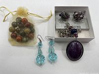 Photo of gemstone marbles, blue crystal drop earrings, purple and silver stud earrings in box, and silver chain necklace with purple stone pendant laid on a white surface.
