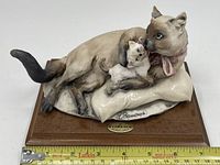 Full view of the porcelain cat figurine with kitten lying on a pillow and wooden base with a measuring tape for scale.