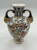 Front view of Tiffany & Co vase showing hand-painted floral and bird decoration with two handles shaped as faces with gold and cobalt blue accents.