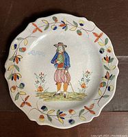 Full view of the round scalloped edge earthenware plate with a hand-painted male figure in historical costume in the center and floral vine border around the rim.