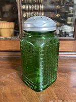 Front and side angled view of antique green glass shaker with metal lid on wooden surface, showing grid texture on glass sides
