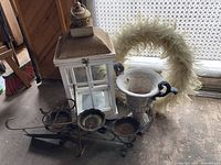 Photo showing a metal lantern, white urn planter, wreath, and candle holder on the floor