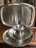 Full view of all four stainless steel dishes including the large rectangular tray at the back and the three smaller dishes in front, one with wooden handles and one bowl with flared rim.