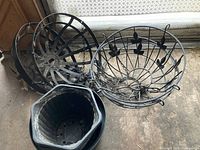 Three black metal garden hanging baskets with decorative leaf accents and chains, and three black plastic pots placed on the floor near a window.