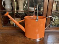 Side view of orange metal watering can showing curved handle and spout with detachable rose nozzle.