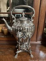 Full view of silver tea kettle on stand showing front detail and decorative feet.
