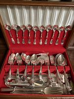 Open box showing organized silver plate cutlery pieces including spoons, forks, knives, and serving utensils in red and white satin-lined wooden box.