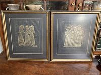 Full view of the pair of framed rubbings showing figures of women in traditional clothing, behind glass in gold frames.