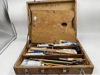 Photo showing artist's case open with wooden palette on lid and assorted paint tubes and brushes inside compartments