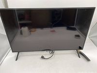 Front view of black Samsung 32 inch TV with power cord attached and remote placed beside it.