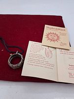 Shepherds Watch personal sundial ring pendant displayed on red fabric pouch next to instruction leaflet.
