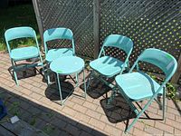 Photo of four light blue powder-coated metal folding chairs and matching round folding table placed outside on a paved surface.