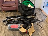 Multiple adjustable camera tripods in black and bronze colors folded and laid on the floor, with a black folding reflector shade and plastic organizer box containing small accessories.
