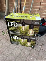 Two unopened boxes of Feit Electric LED string lights, 48ft each, displayed outside against a brick wall and gardening tools.