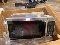 Microwave in cardboard box showing front and top sides, stainless steel surface, control panel and handle.