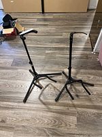 Two black adjustable musical instrument stands with tripod bases and padded support arms on a wood floor.