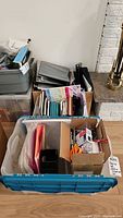 Top view of three plastic bins filled with assorted office supplies including folders, binders, papers, and stationery items