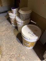 Five containers of paint remnants stacked on a concrete floor in a corner, shows overall lot quantity and container types.