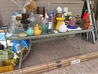 Full view of the patio table with glass top covered with various household items, showing table shape, color, and condition.