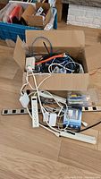 Box containing tangled cables, power bars, and a plastic box with hand tools or small parts, placed on floor.