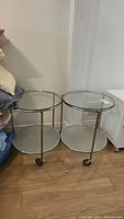 Pair of round side tables with metal frames, clear glass top shelves, frosted glass lower shelves on caster wheels, shown side by side on wooden floor.