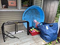 Full lot showing hockey nets, wading pool, beach toys in basket, blue bag, and red sled on wooden deck by house