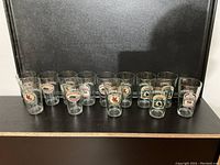 Front view of 13 clear beer glasses with various printed logos aligned in a row against a black background.